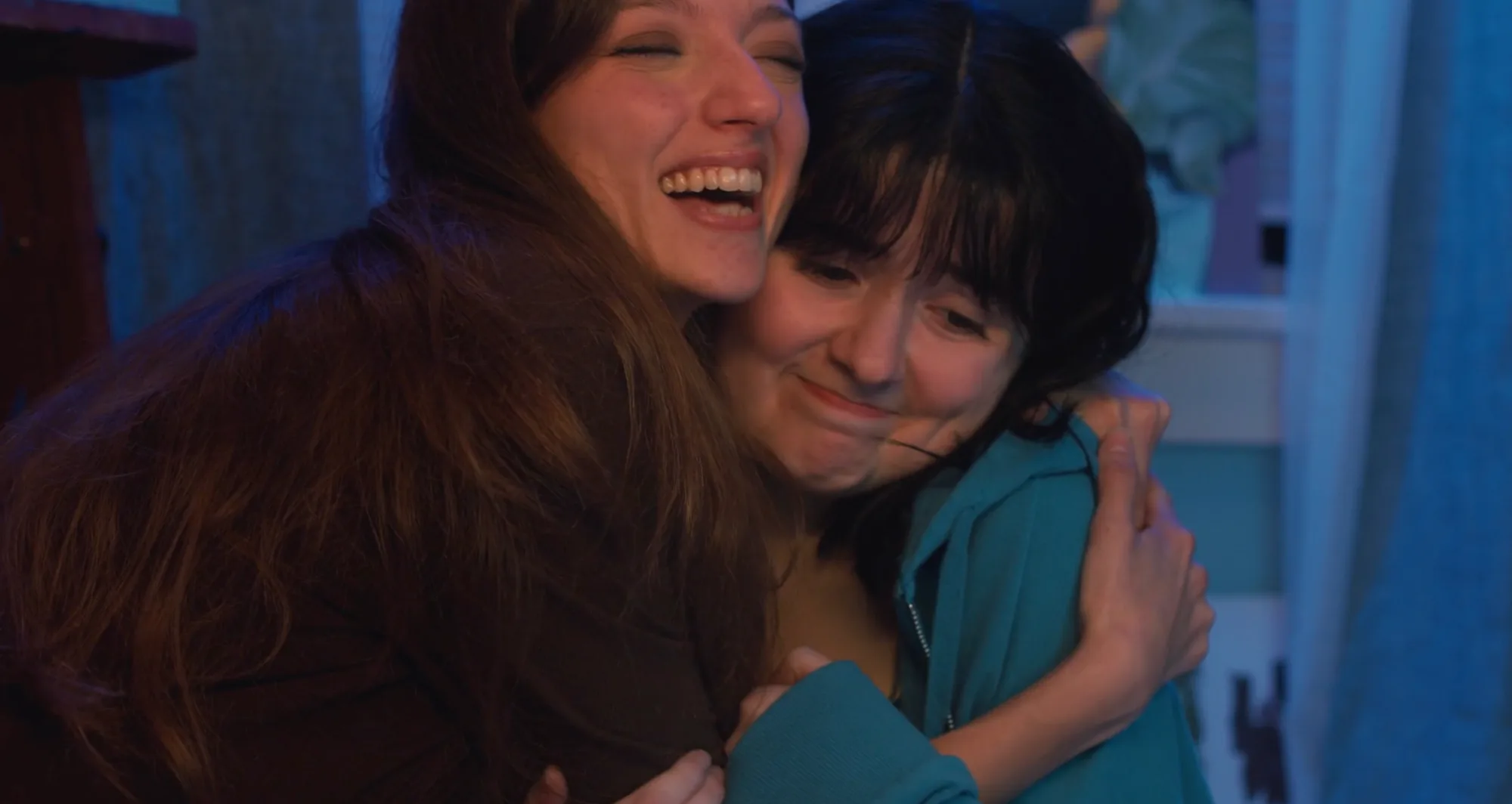 Two teenage girls hugging and laughing in a blue-lit bedroom.