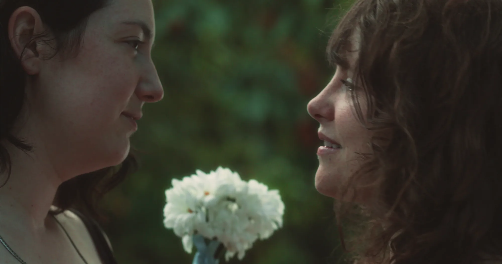 Two women face to face outdoors, a small bouquet of daisies between them.